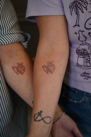 Close-up of two forearms showing matching minimalist fine-line heart tattoos with tiny bows on the inner arms; one arm also features an anchor‑infinity tattoo and beaded bracelet, casual striped and graphic tees visible.