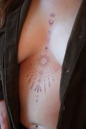 Close-up of a person’s torso featuring a minimalist celestial sternum tattoo with crescent moon, sunburst and dotted rays, open brown ribbed cardigan and navel piercing.