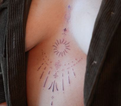 Close-up of a person’s torso featuring a minimalist celestial sternum tattoo with crescent moon, sunburst and dotted rays, open brown ribbed cardigan and navel piercing.