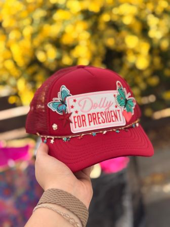 Hand holding a red trucker hat with a 'Dolly for President' embroidered patch, turquoise butterfly appliqués and star charm trim against a blurred yellow floral outdoor background.