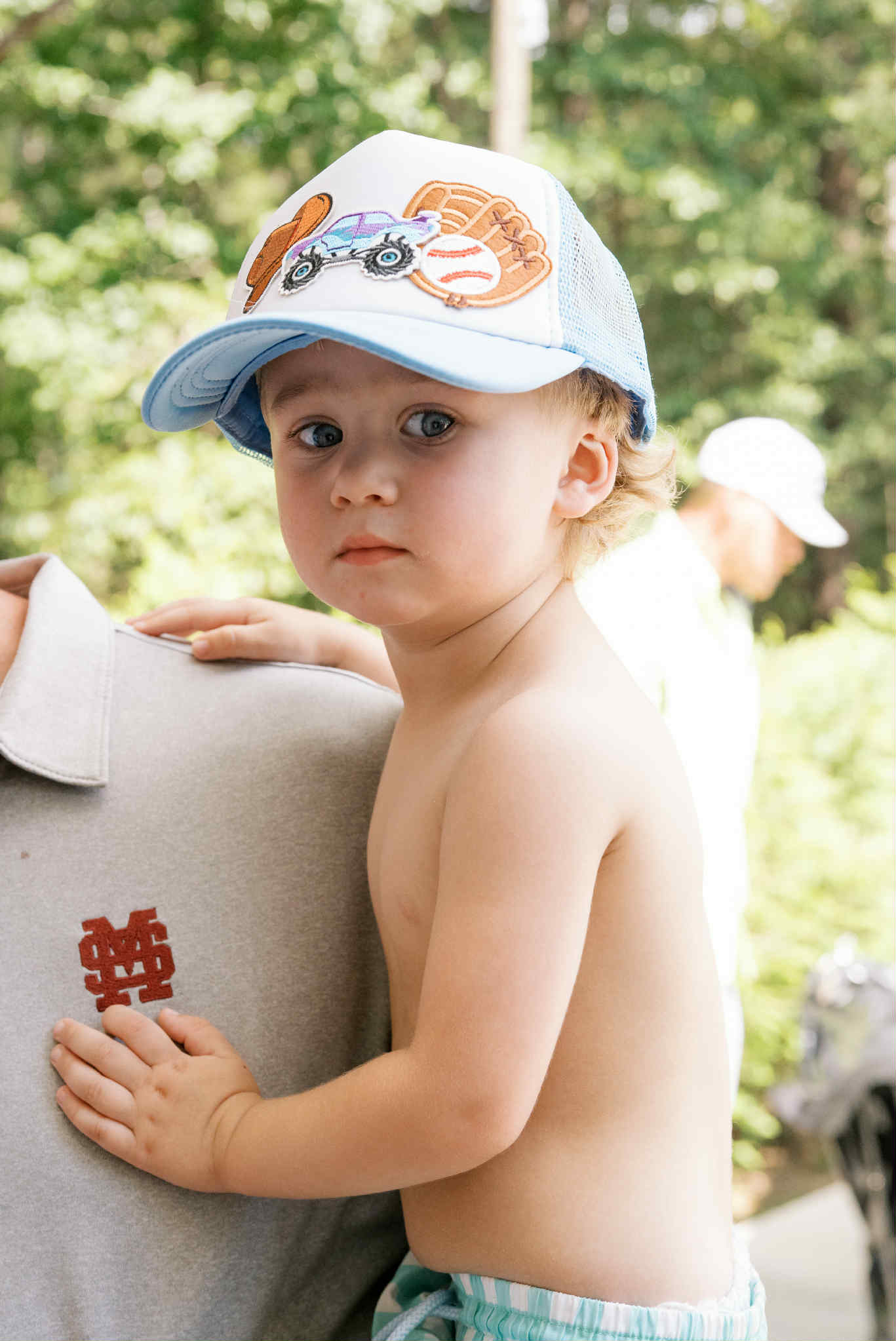 Shirtless toddler wearing a blue trucker cap with a toy car and baseball patches, leaning on an adult in a sunlit outdoor setting