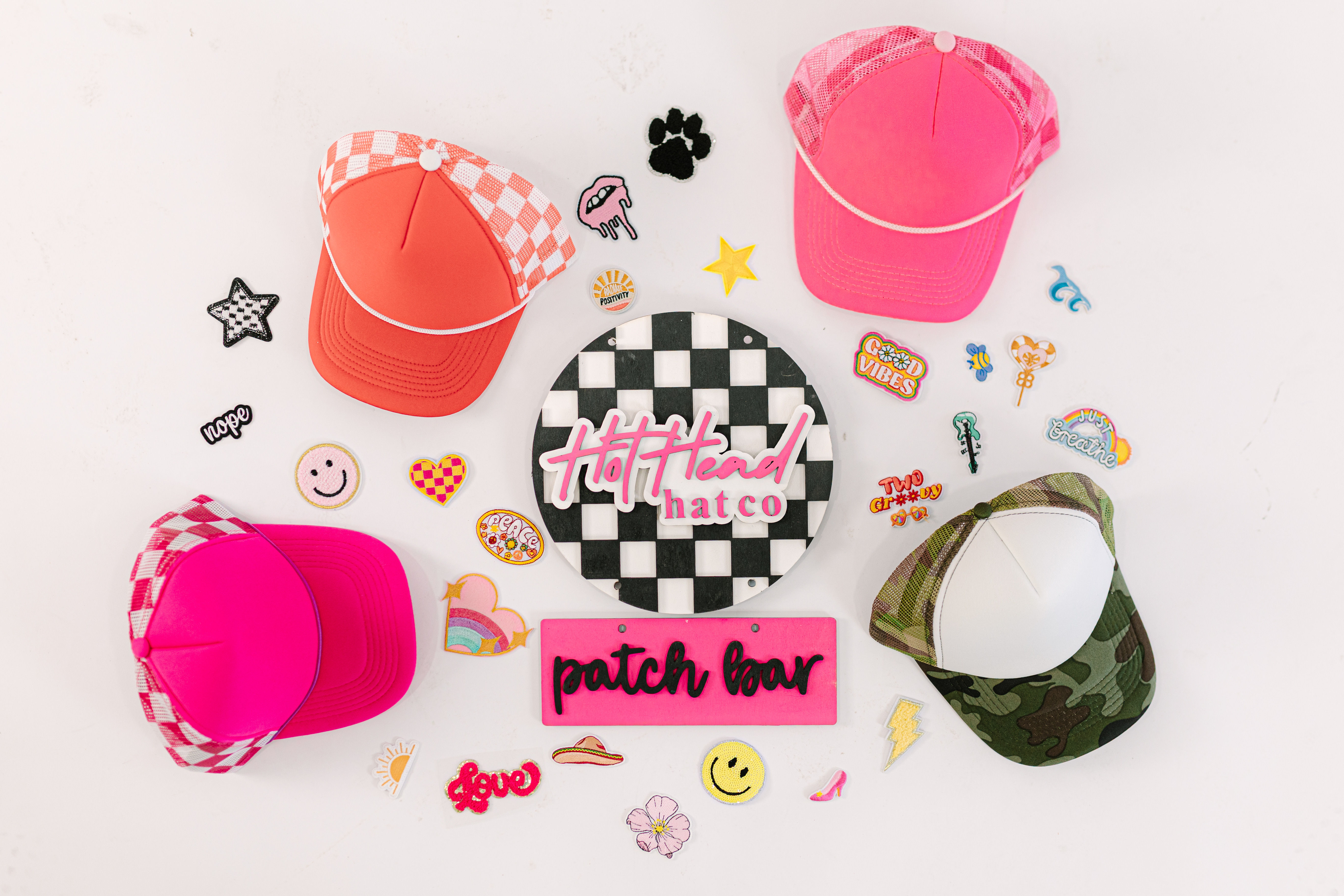 Bright pink, coral and camo trucker caps arranged with colorful embroidered patches around a black-and-white checkerboard sign on a white background