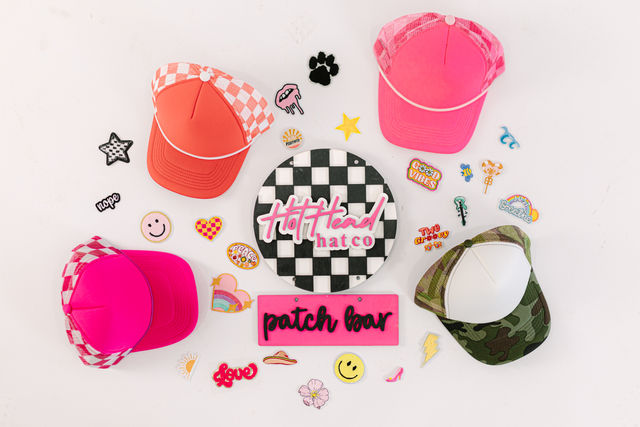 Bright pink, coral and camo trucker caps arranged with colorful embroidered patches around a black-and-white checkerboard sign on a white background