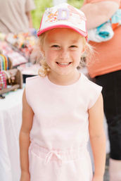 Beaming young girl wearing a pink embellished baseball cap and matching sleeveless outfit at an outdoor market stall