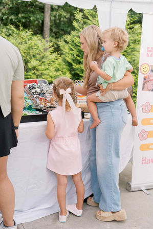Parent holding a toddler while a young girl with a pink bow browses colorful stickers and patches at an outdoor craft fair booth under a white tent with leafy greenery behind them.