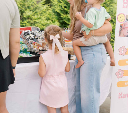 Parent holding a toddler while a young girl with a pink bow browses colorful stickers and patches at an outdoor craft fair booth under a white tent with leafy greenery behind them.