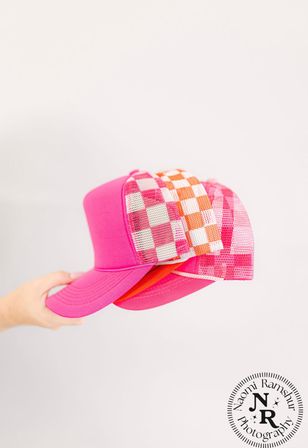 Bright pink trucker hats with orange-and-white checkered mesh panels stacked and held by a hand against a clean white background