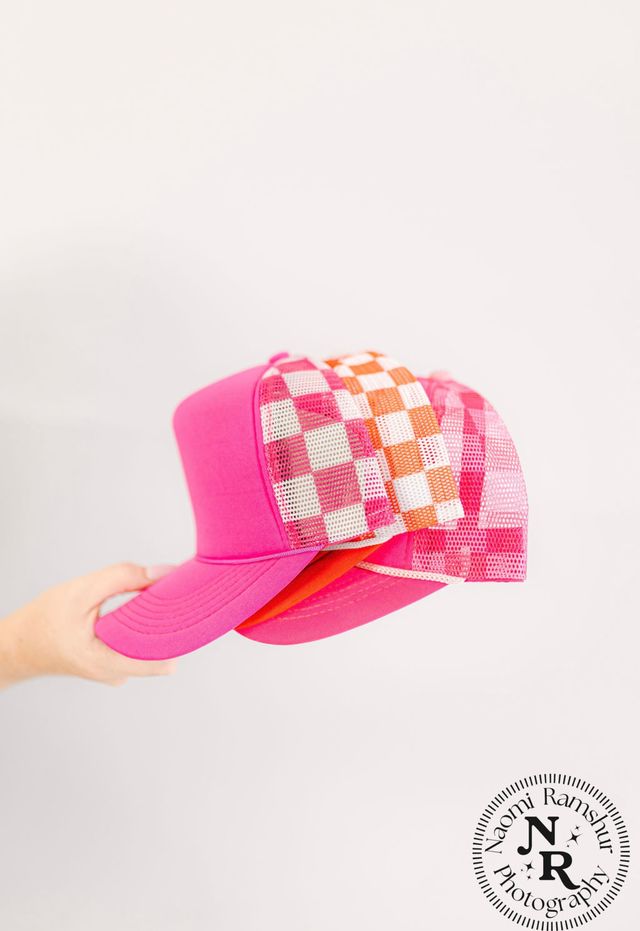 Bright pink trucker hats with orange-and-white checkered mesh panels stacked and held by a hand against a clean white background
