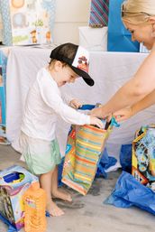 Toddler in a flame trucker hat eagerly unwrapping a colorful gift bag while an adult helps, surrounded by wrapped presents and blue tissue paper at a kids' party.