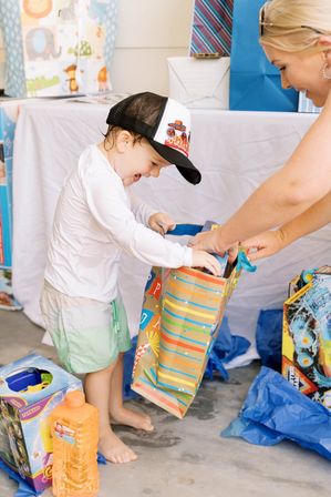 Toddler in a flame trucker hat eagerly unwrapping a colorful gift bag while an adult helps, surrounded by wrapped presents and blue tissue paper at a kids' party.