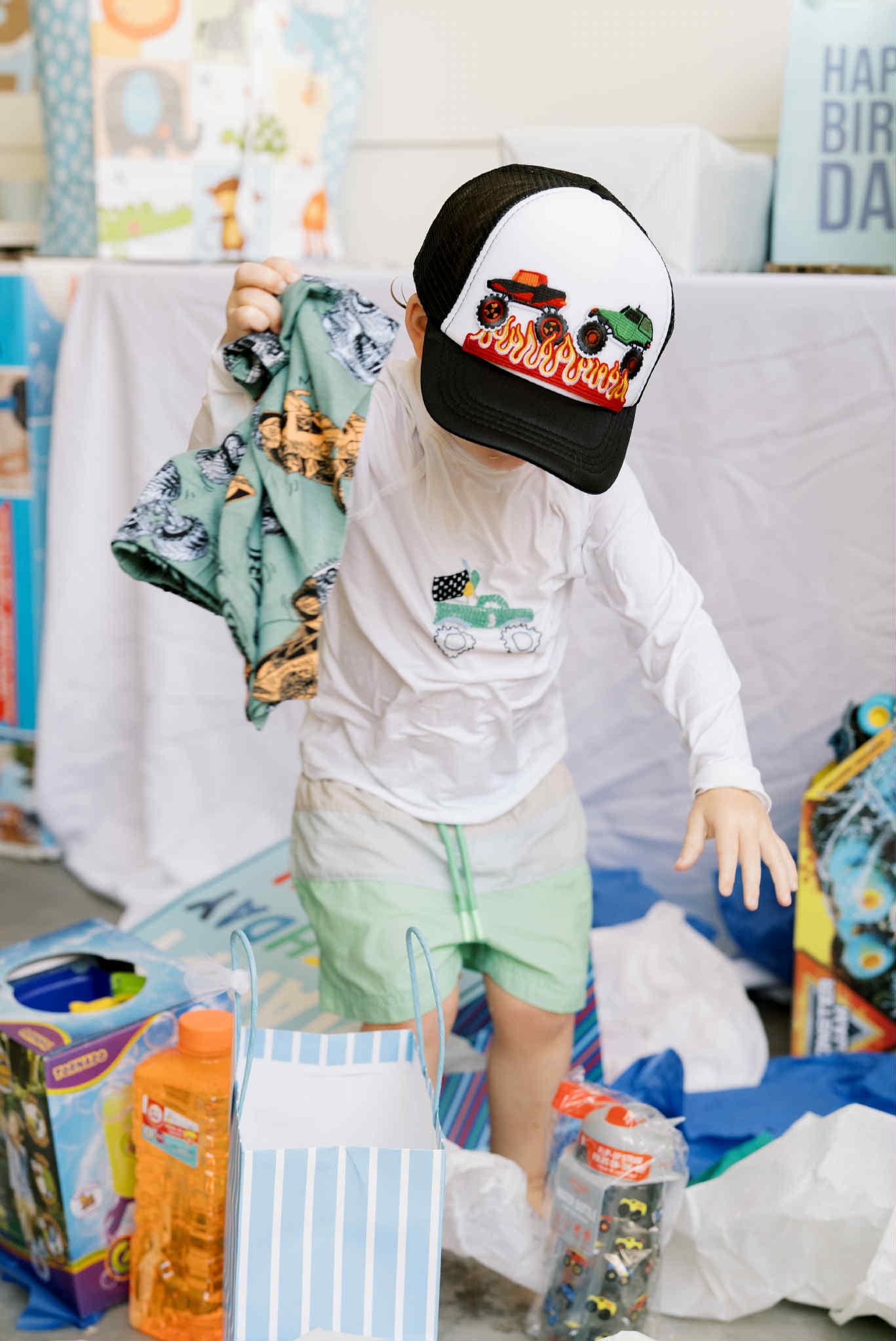Young child at a birthday party wearing a monster-truck baseball cap with flames, holding a patterned blanket amid opened presents, toy packaging and blue-striped gift bags.