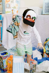 Young child at a birthday party wearing a monster-truck baseball cap with flames, holding a patterned blanket amid opened presents, toy packaging and blue-striped gift bags.