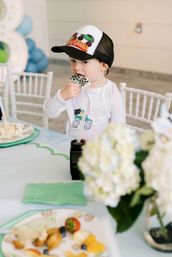 Toddler in a truck baseball cap biting a checkered cookie at a blue-themed indoor kids' birthday party table with plates and balloons