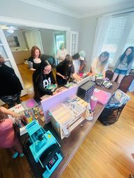 At-home craft workshop in a bright living room with a group of women gathered around a long table browsing DIY jewelry and craft supplies — beads, storage boxes, scissors and tools spread across the workspace.