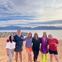 Six friends posing arm‑in‑arm on a sandy alpine lake beach with a rocky breakwater, snow‑capped mountains and cloudy sky; two hold red cups and one wears a bright purple robe with neon leggings.