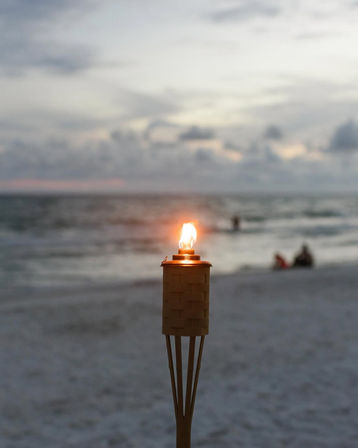 Glowing tiki torch on a sandy beach at dusk, ocean waves and a soft sunset sky in the background.