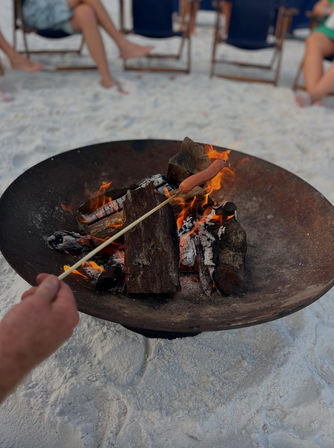 Hand holding a skewer roasting a hot dog over a wood fire in a metal beach fire pit on white sand with folding chairs nearby