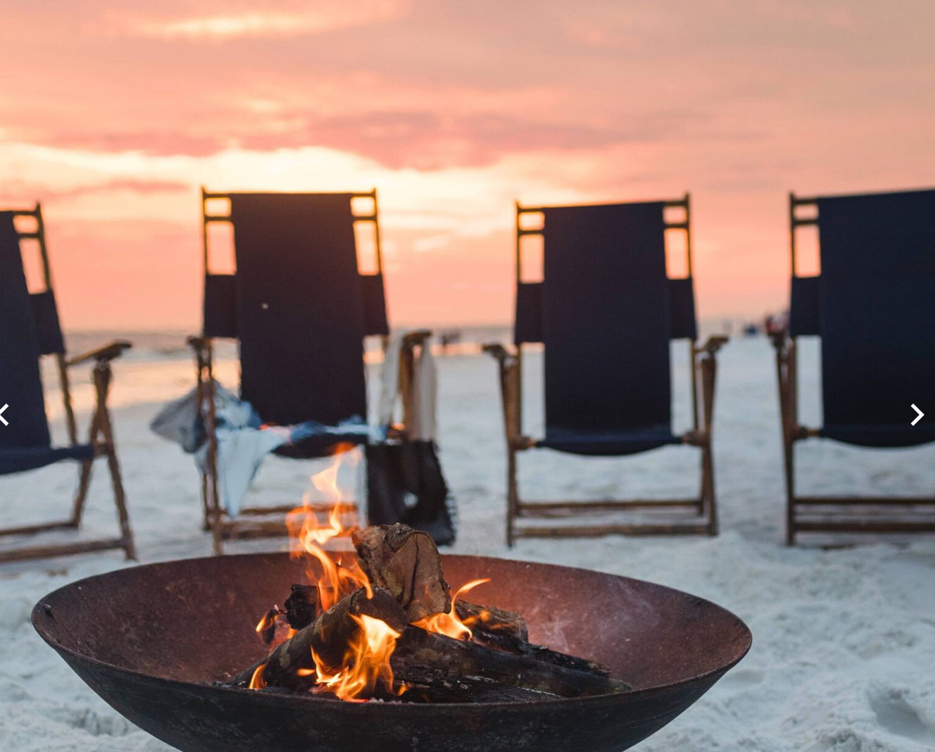 Cozy beach bonfire in a metal bowl on white sand with empty navy lounge chairs facing the ocean under a pink sunset