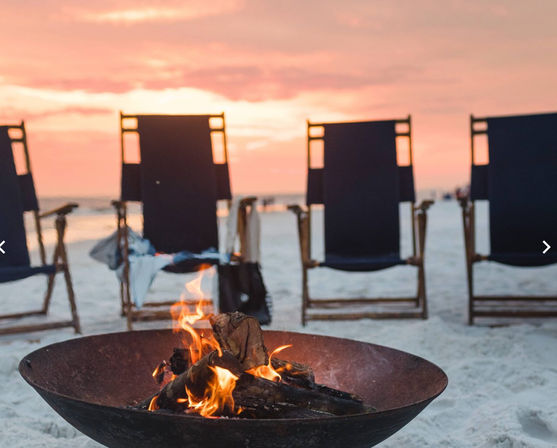 Cozy beach bonfire in a metal bowl on white sand with empty navy lounge chairs facing the ocean under a pink sunset