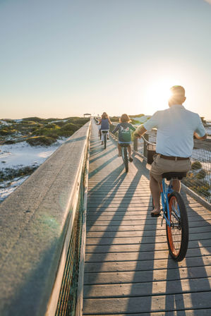 Three cyclists riding on a coastal beach boardwalk at sunset, long shadows stretching over sand dunes.