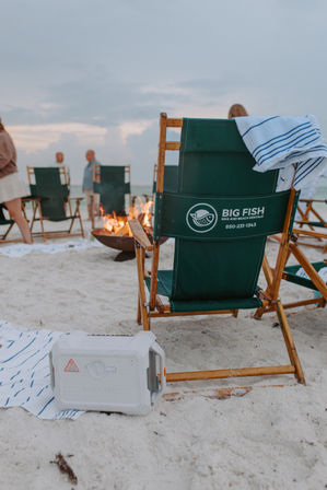 Green wooden beach chair with striped towel on white sand beside a cooler and a small bonfire, blurred beachgoers and ocean at sunset — relaxing coastal scene.
