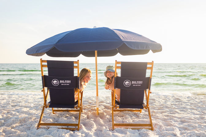 Two children smiling from matching navy beach chairs under a large navy umbrella on a sunny white‑sand beach with calm green ocean waves and bright sky in the background.