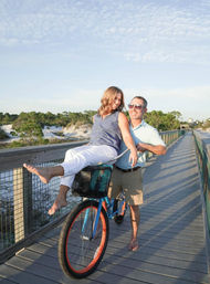 Playful couple on a sunny beach boardwalk — woman sitting barefoot in a bicycle’s front basket with legs out, man pushing and smiling, coastal dunes and blue sky in the background.