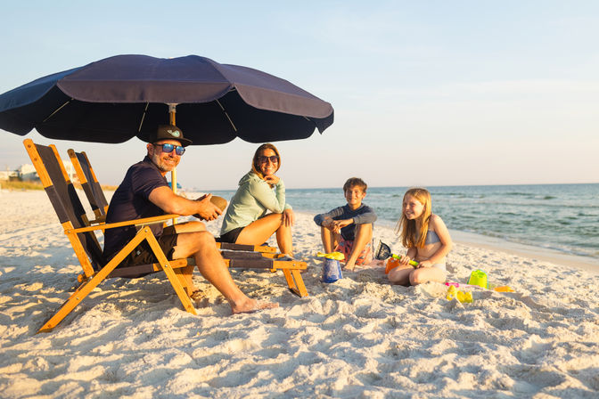 Smiling family of four enjoying a sandy seaside at golden hour — parents lounging in wooden chairs under a navy umbrella while two kids build sandcastles with colorful toys by the ocean
