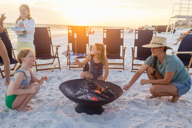 Kids and an adult roasting marshmallows over a portable fire pit on a white-sand beach at sunset with folding chairs in the background.