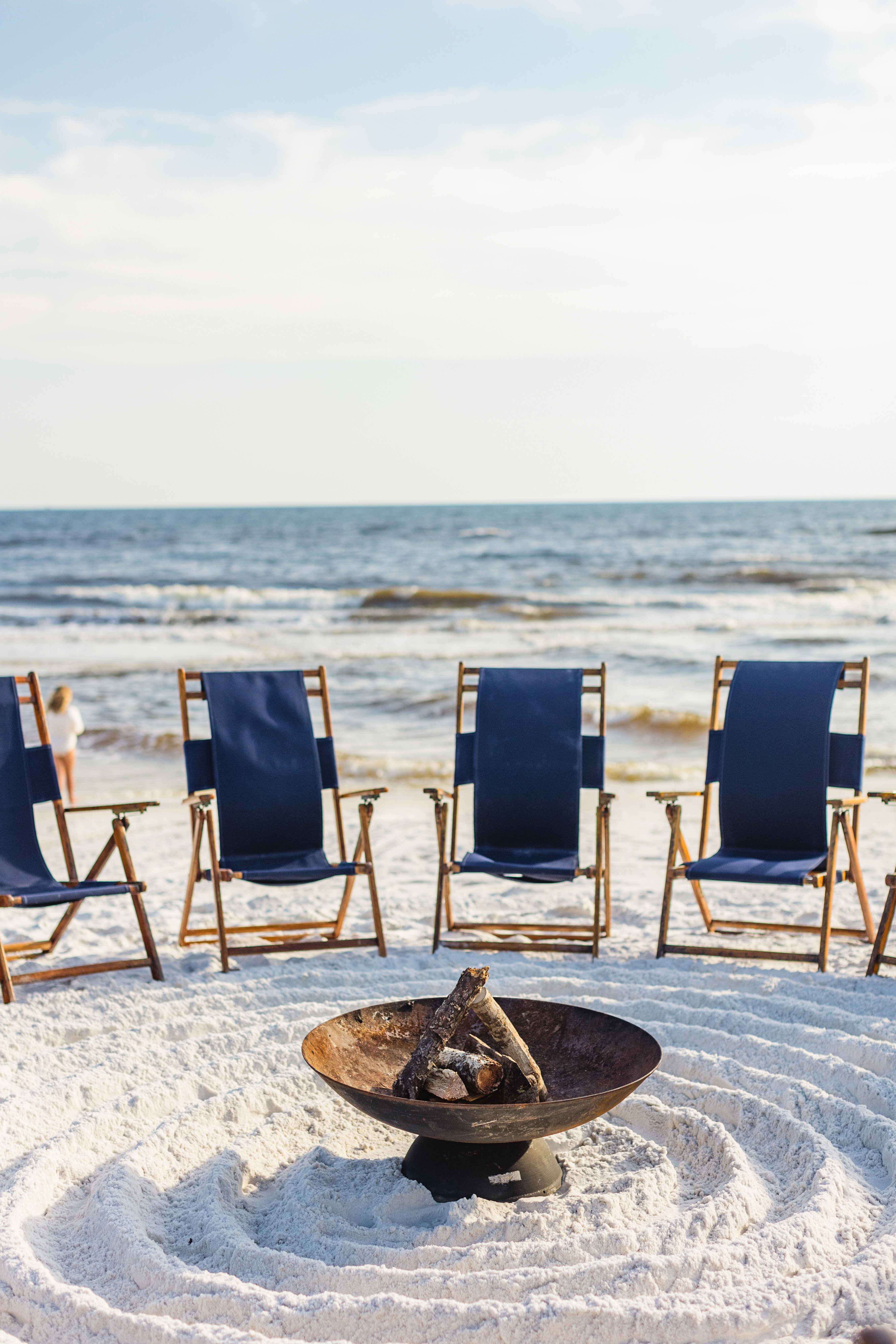 Metal fire pit with stacked logs in a raked white-sand circle, navy beach chairs lined up facing gentle ocean waves under a pale sky