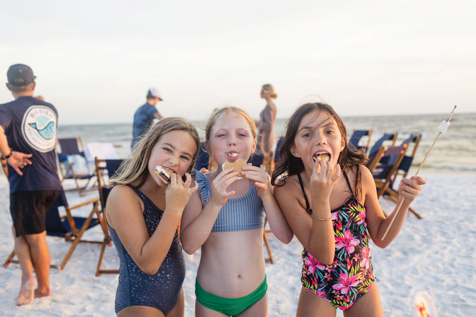 Three girls in colorful swimsuits eating s'mores on a sandy ocean beach at sunset, with folding chairs and people in the background