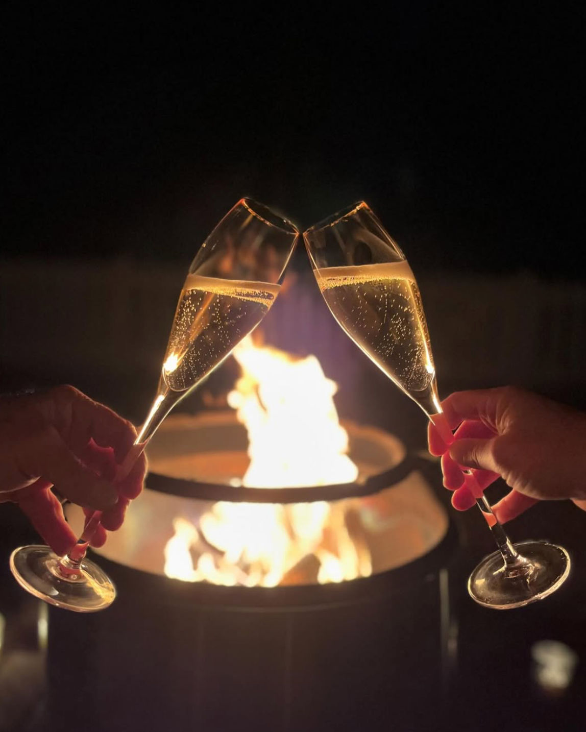 Two bubbling champagne flutes clinking above a warm outdoor fire pit at night — hands toasting in a cozy celebration.