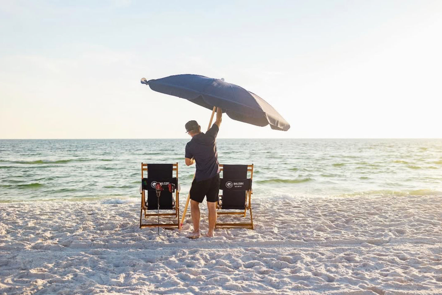Man setting a large navy beach umbrella between two wooden lounge chairs on a sunlit sandy beach by the calm ocean at sunset