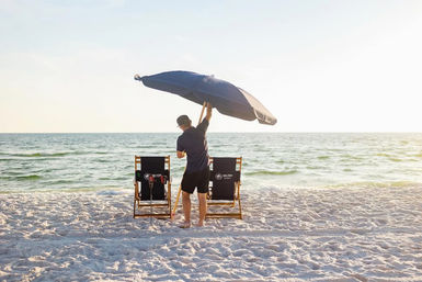Man setting a large navy beach umbrella between two wooden lounge chairs on a sunlit sandy beach by the calm ocean at sunset