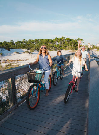 Smiling family riding colorful cruiser bikes on a sunny coastal boardwalk past sand dunes and pine trees