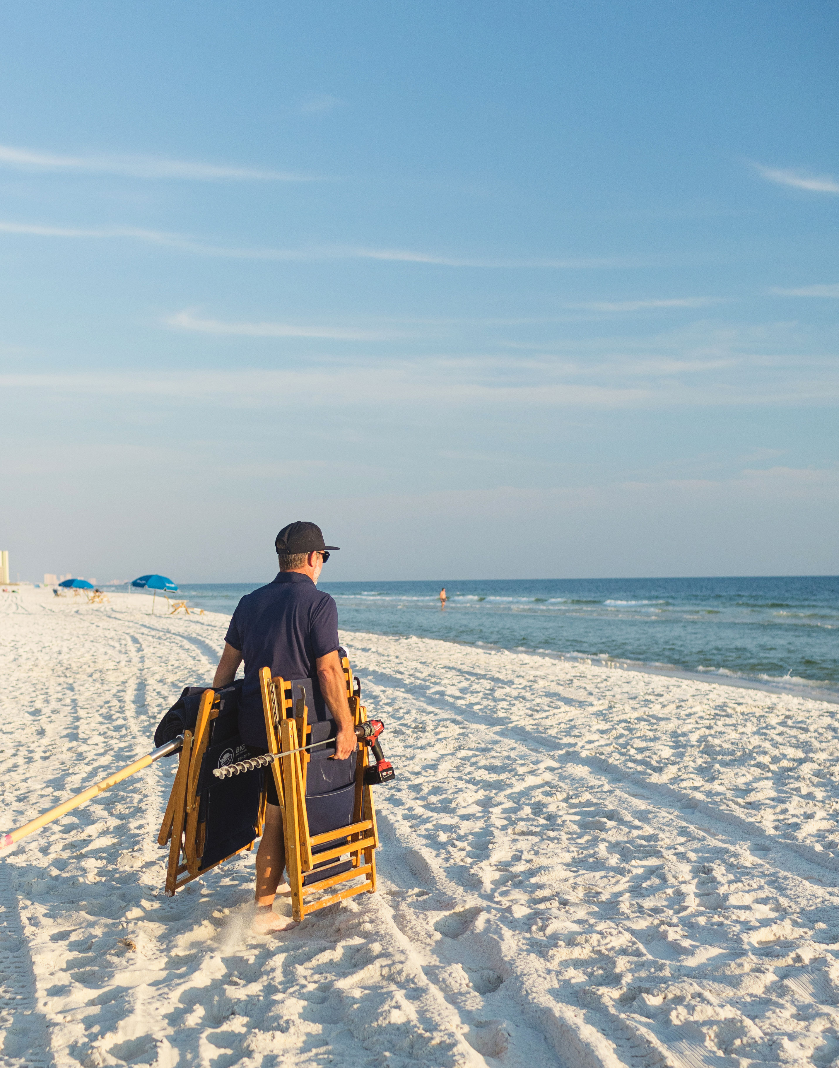 Beach-goer carrying wooden folding chairs and an umbrella pole across a sunlit white-sand shore toward a calm blue ocean under a clear sky