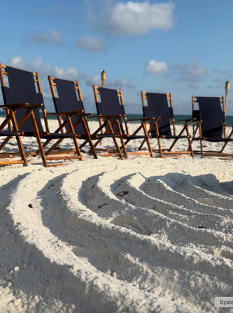 Row of wooden beach chairs with navy fabric on white sandy beach, raked sand patterns, turquoise ocean and blue sky with tiki torches