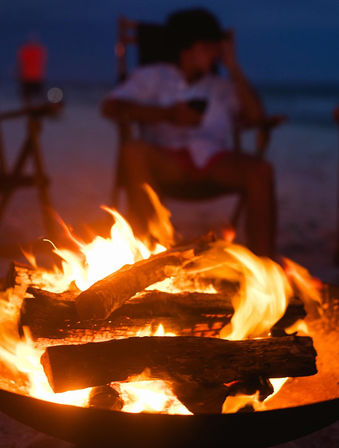 Cozy beach bonfire at dusk — glowing logs crackling in a fire pit with a blurred person relaxing in a chair by the shoreline