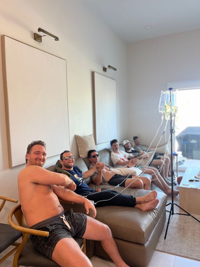 Group of men lounging on a living-room sofa, smiling while IV fluid bags and tubing hang from a stand by a sunlit window, neutral wall art and modern decor in the background.