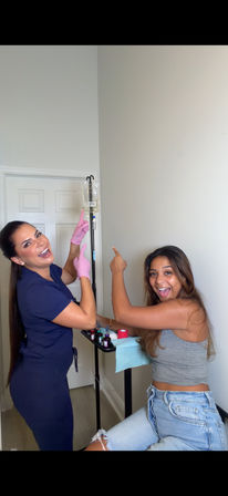 Cheerful clinician in scrubs with pink gloves and a smiling patient pointing at a hanging IV drip in a compact wellness clinic treatment room with a small medical tray.