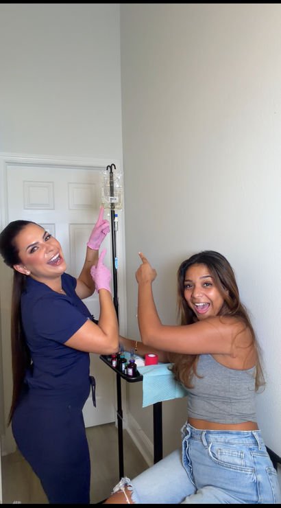 Cheerful clinician in scrubs with pink gloves and a smiling patient pointing at a hanging IV drip in a compact wellness clinic treatment room with a small medical tray.
