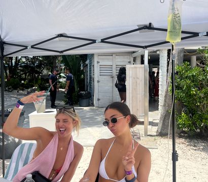 Two women in bikinis laughing and posing under a white canopy at a beachside outdoor booth, an IV drip bag hangs from a pole beside the table with bottled water, sandy path and tropical foliage in the background.