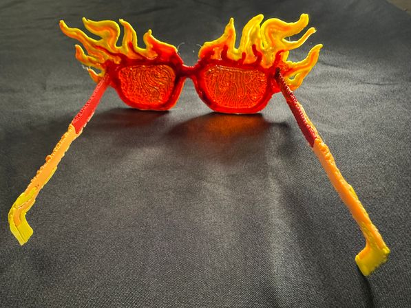 Bright red-and-yellow flame-shaped novelty sunglasses with wavy fiery frames and long temples on black fabric — playful party or festival accessory.