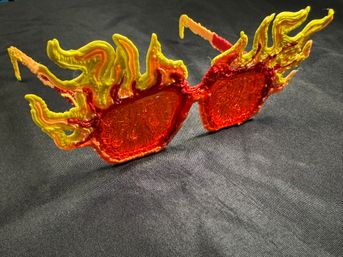 Novelty flame sunglasses with translucent red-orange lenses and sculpted yellow-orange flame-shaped plastic frames resting on black fabric