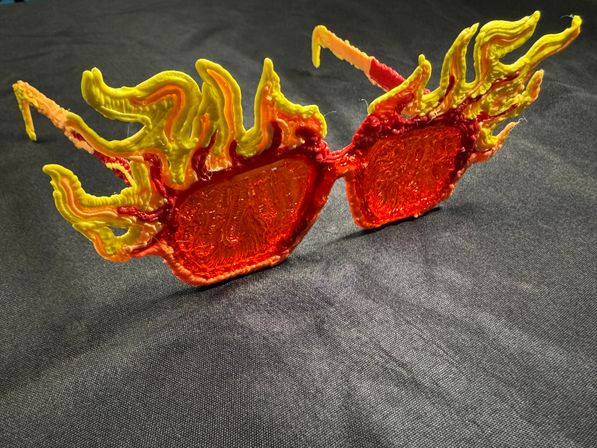 Novelty flame sunglasses with translucent red-orange lenses and sculpted yellow-orange flame-shaped plastic frames resting on black fabric
