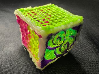 Close-up of a playful handmade neon 3D-pen cube sculpture with a green lattice top, bright pink and yellow lattice sides, and an iridescent green swirl panel framed in purple, sitting on dark textured fabric.
