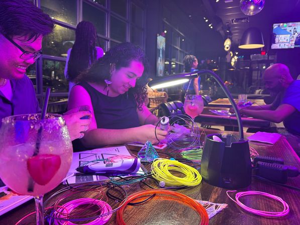 Hands-on craft night at a bar: people working with a 3D-printing pen and bright coiled filament under purple neon lights, cocktails on the table and a desk lamp illuminating a modern restaurant interior with large windows and pendant lights.