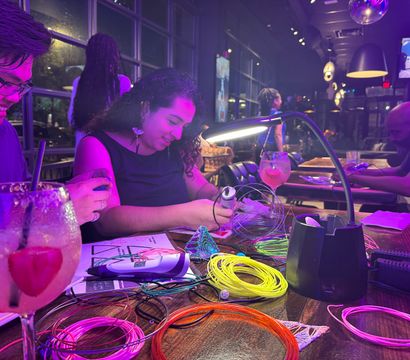 Hands-on craft night at a bar: people working with a 3D-printing pen and bright coiled filament under purple neon lights, cocktails on the table and a desk lamp illuminating a modern restaurant interior with large windows and pendant lights.