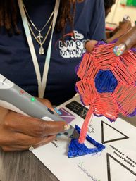 Close-up of hands using a 3D printing pen to create a vibrant filament flower sculpture in pink, blue and purple on a makerspace table, decorated nails and glowing pen tip visible.