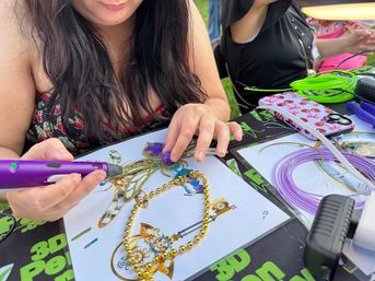Hands using a purple 3D printing pen to add purple filament to a steampunk insect design on paper at an outdoor craft table with gold beads, coils of filament and crafting tools.
